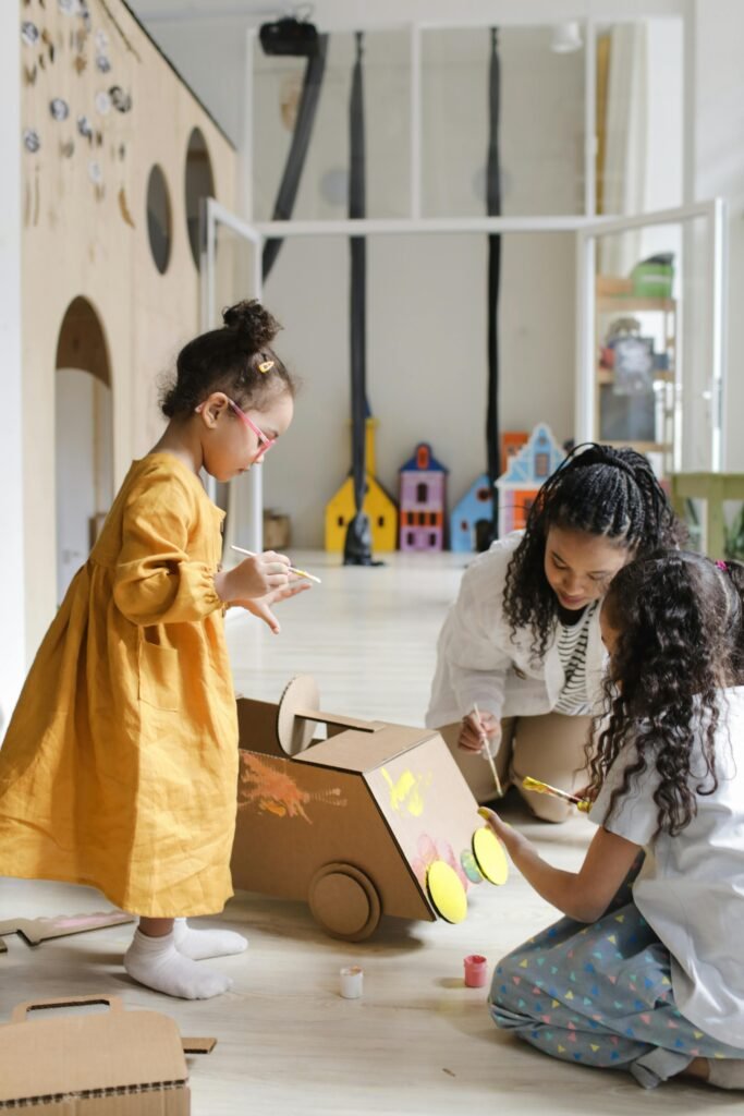 pexels-photo-8540374-8540374 Two young girls painting a cardboard box car indoors, guided by a caregiver in a nurturing environment.