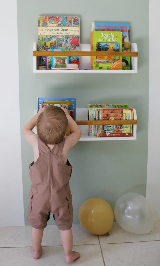 pexels-photo-36455220-36455220 A toddler stands reaching for books on a wall shelf, surrounded by colorful balloons.
